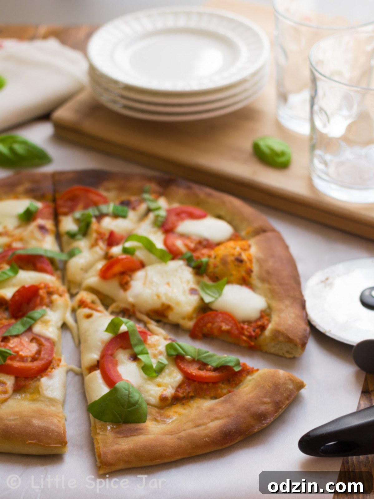 Fresh basil leaves artfully placed on a sliced Margherita pizza, resting on a white cutting board, with plates and cups softly blurred in the background.