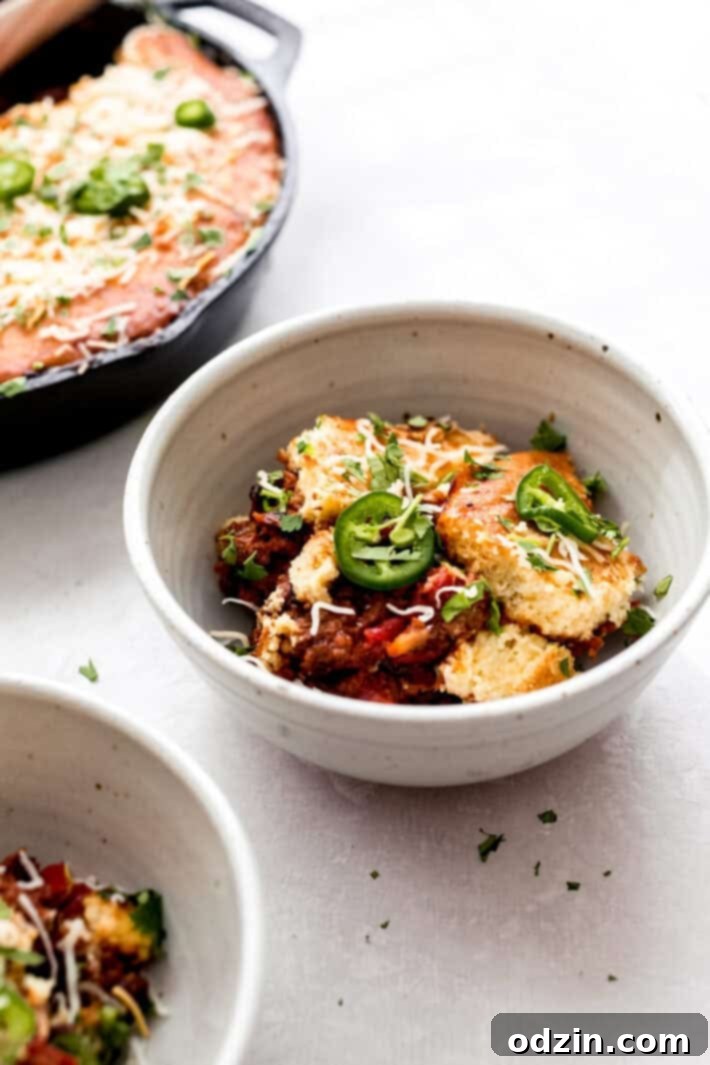 Jalapeño Cheddar Cornbread-Capped Skillet Chili 2 bowl with prepared skillet chili and broken cornbread crust on grey surface surrounded by skillet and another bowl
