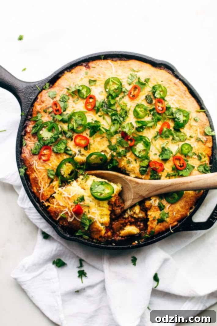Jalapeño Cheddar Cornbread-Capped Skillet Chili 3 skillet with chili topped with cornbread with a wooden spoon lifting a piece out on white surface with white tea towel