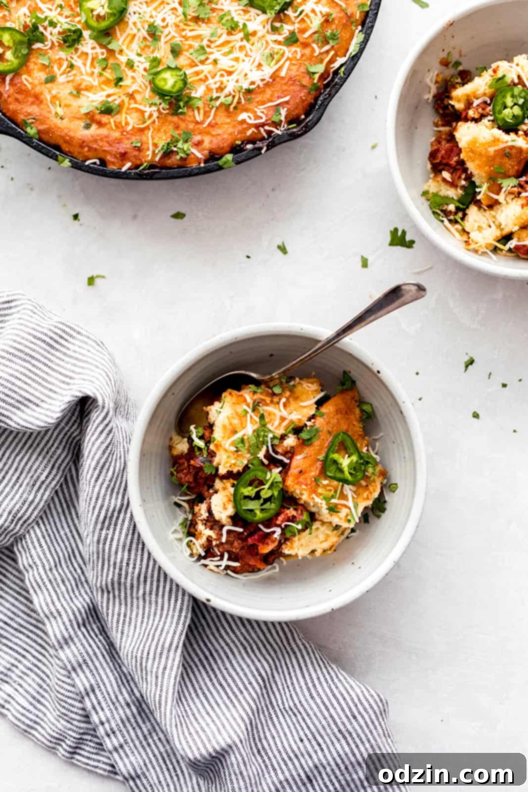 Jalapeño Cheddar Cornbread-Capped Skillet Chili 9 food scene on grey surface with bowls of chili and skillet with striped tea towel