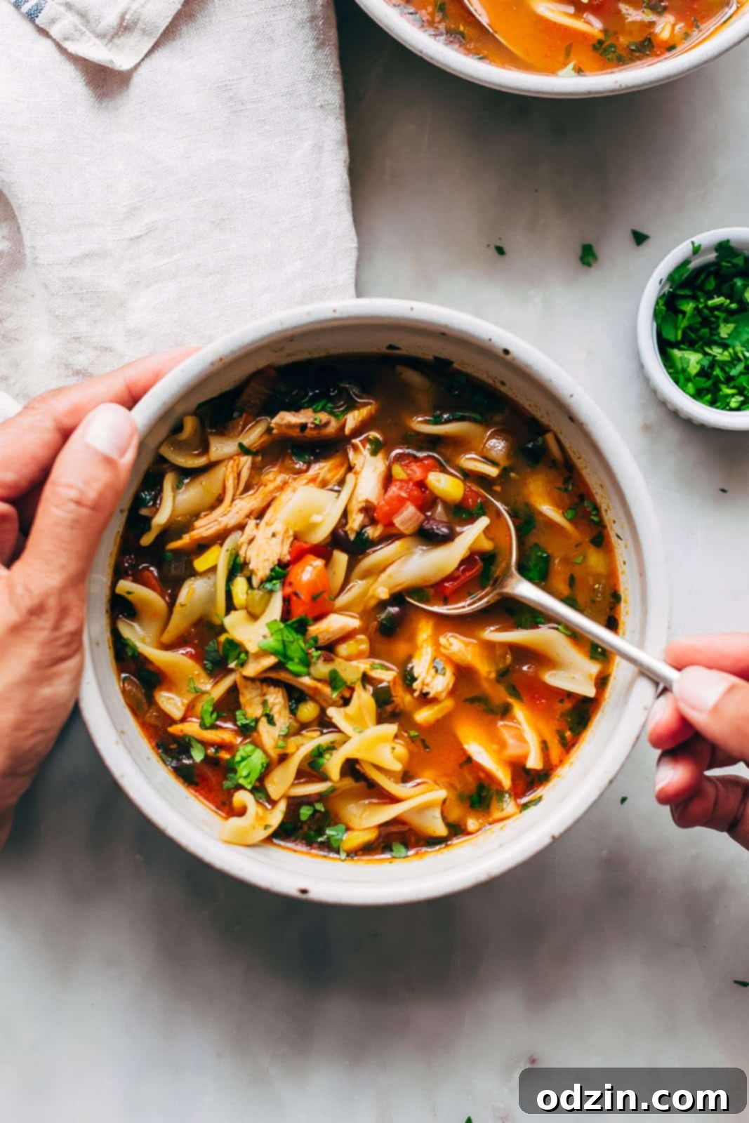 A spoon resting in a bowl of Mexican chicken noodle soup, held by a hand, against a white marble background, highlighting the soup's comforting appeal.
