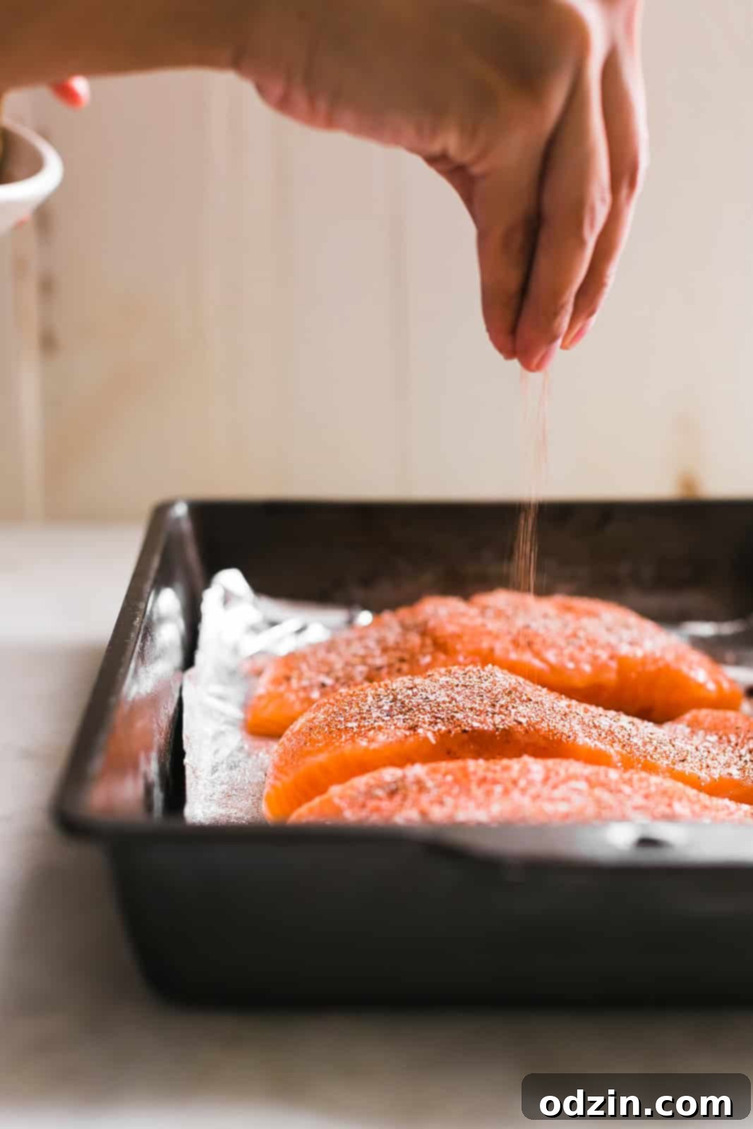 Sprinkling homemade seasoning rub on raw salmon fillets arranged in a metal baking dish, prior to cooking.