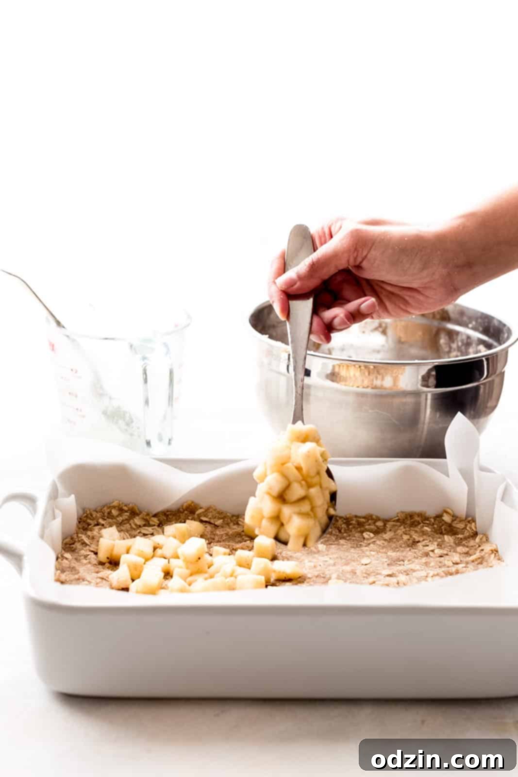 Golden Apple Crumble Bars 7 hand with spoon sprinkling diced apples over crust in white baking dish