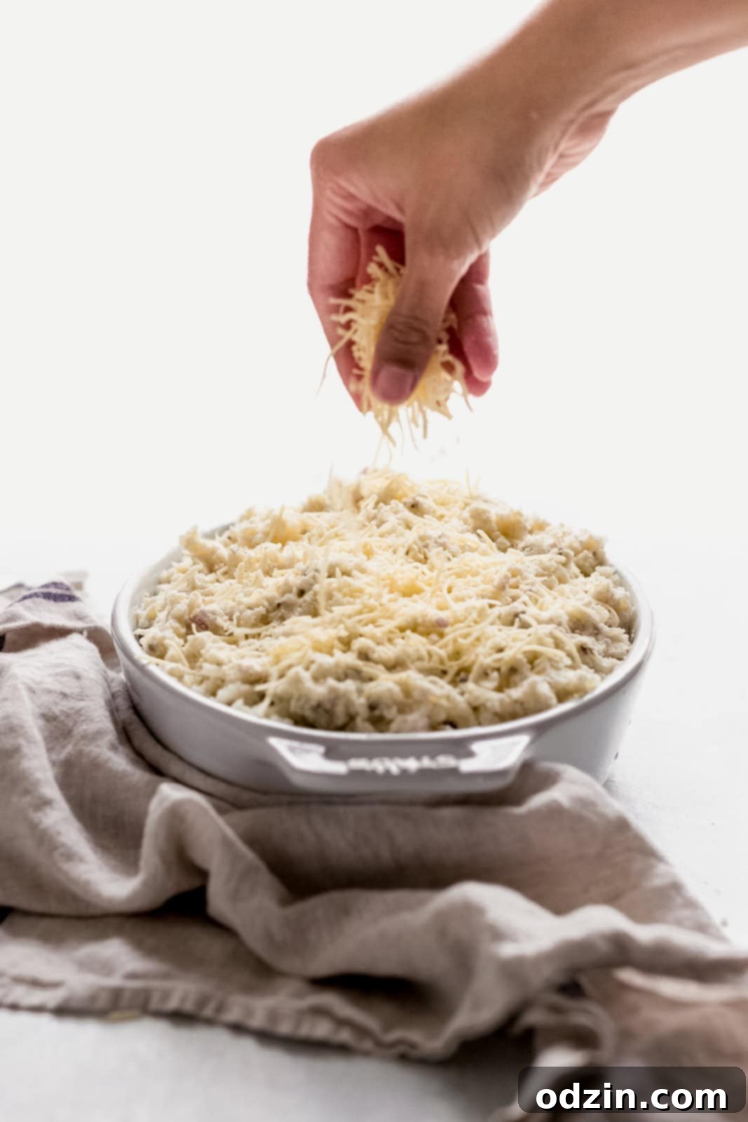 A hand gently sprinkles shredded cheese over a casserole dish filled with fluffy, seasoned potato mixture, ready for baking.