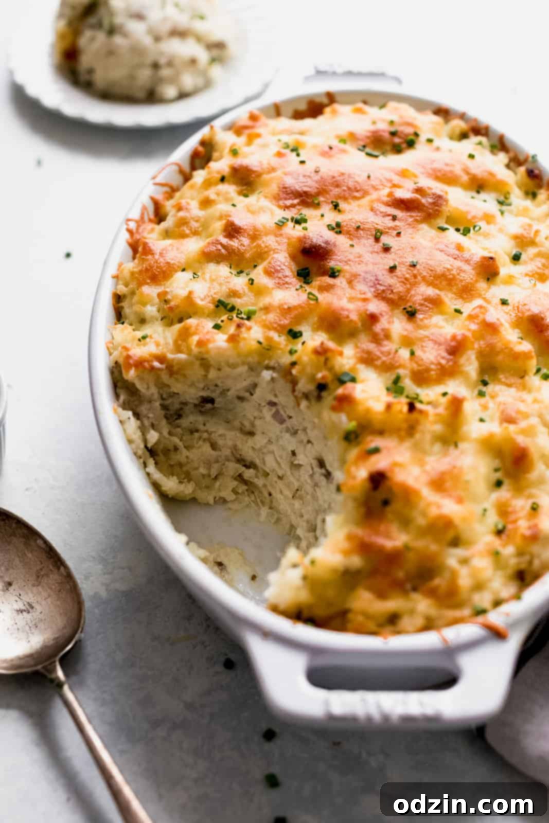 A serving spoon lifting a scoop of bubbly, cheese-topped baked potatoes from a white casserole dish, showcasing its rich texture and melted cheese topping.