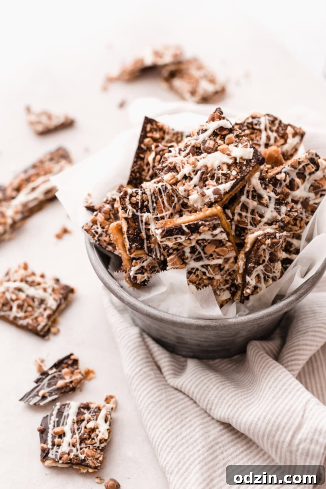 bucket of saltine toffee bark on striped towel surrounded by pieces of toffee on grey surface