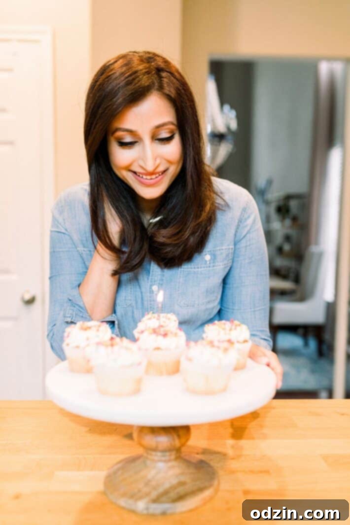 Author standing proudly next to a cake stand adorned with six beautifully decorated cupcakes, one featuring a lit candle, marking the blog's 5th anniversary.