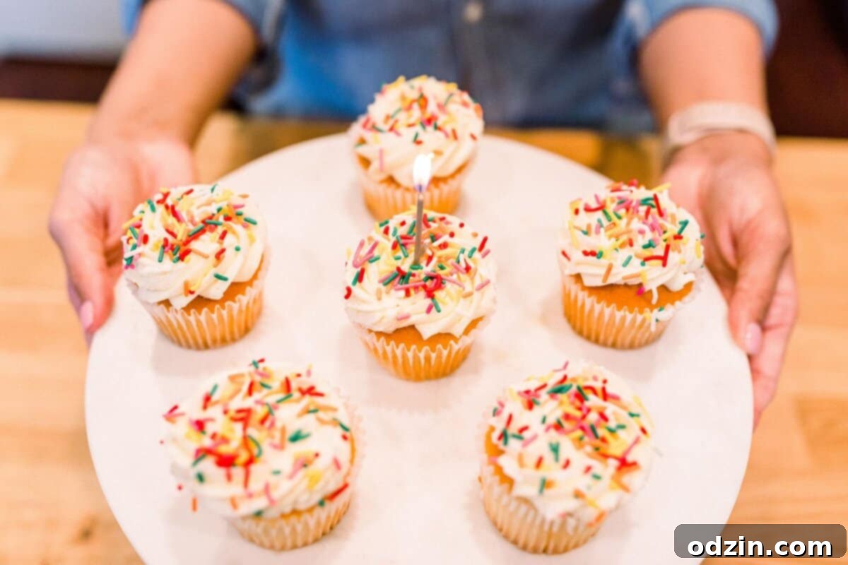 Close-up shot of hands gently holding a cake stand laden with festive cupcakes, one proudly displaying a lit anniversary candle.