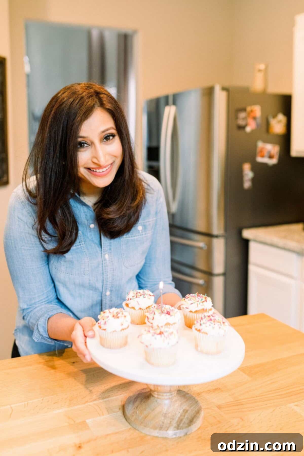 The author smiling warmly at the camera, holding a cake stand adorned with a variety of colorful and tempting cupcakes.