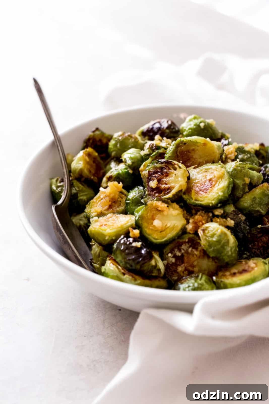 Side view of a white bowl overflowing with golden-brown roasted Brussels sprouts coated in garlic butter, with a serving spoon nestled in, placed on a grey surface next to a white tea towel.