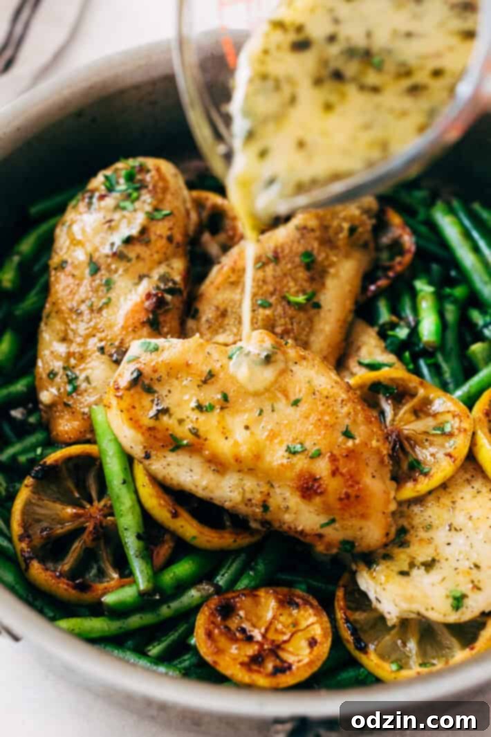 Pouring bright lemon butter sauce over perfectly seared chicken breasts in a skillet with fresh lemon slices and green beans