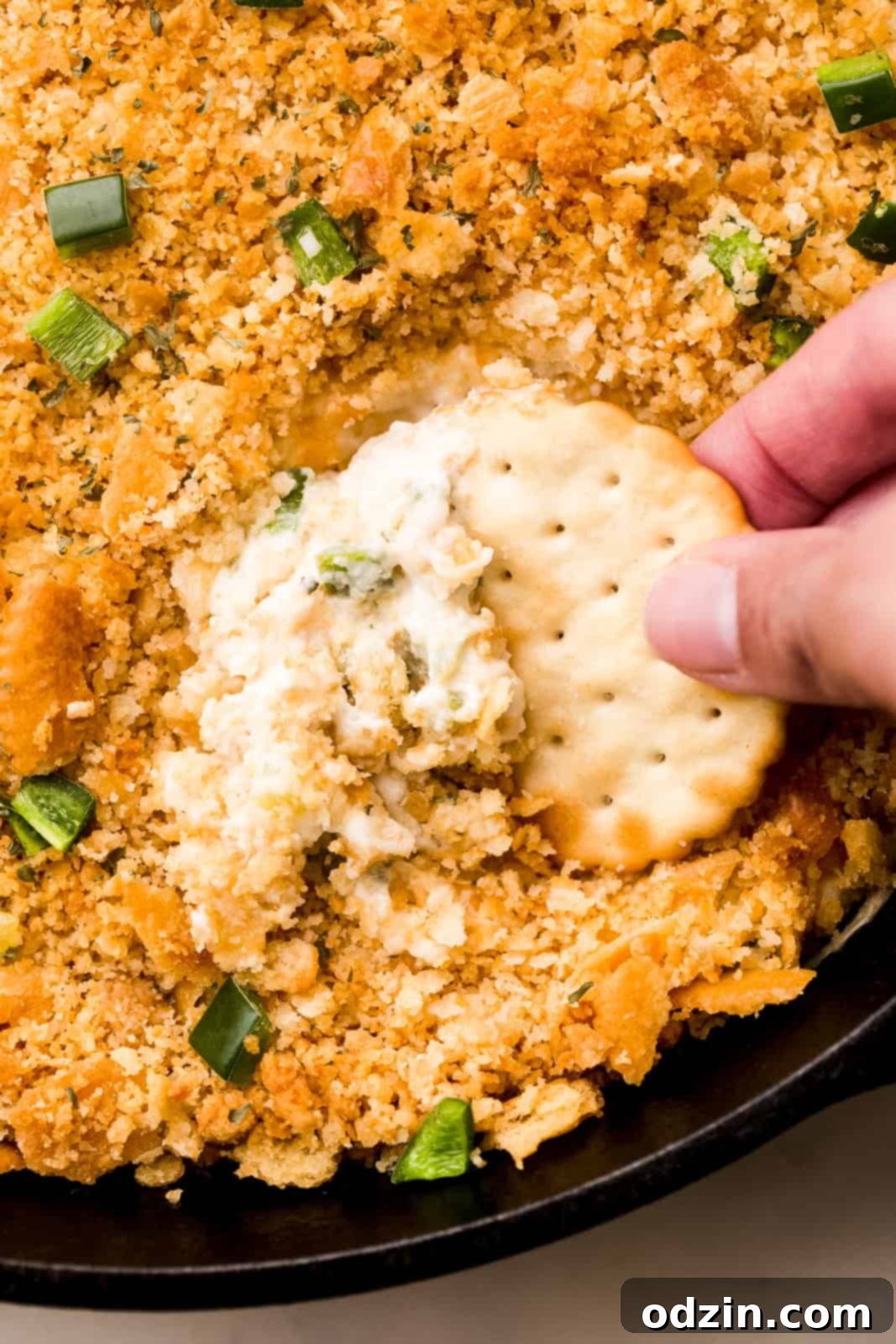 Close-up of fingers holding a cracker lifting creamy jalapeño popper dip from a skillet