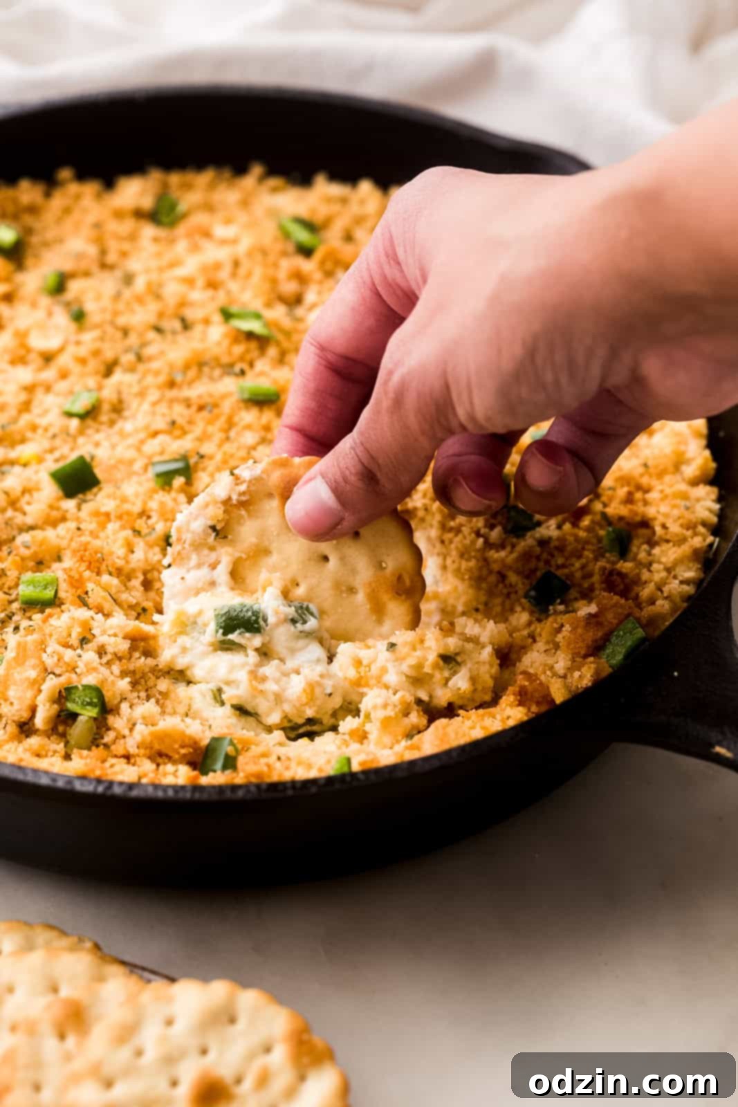 Hand dipping a cracker into a bubbly, warm jalapeño popper dip in a cast iron skillet