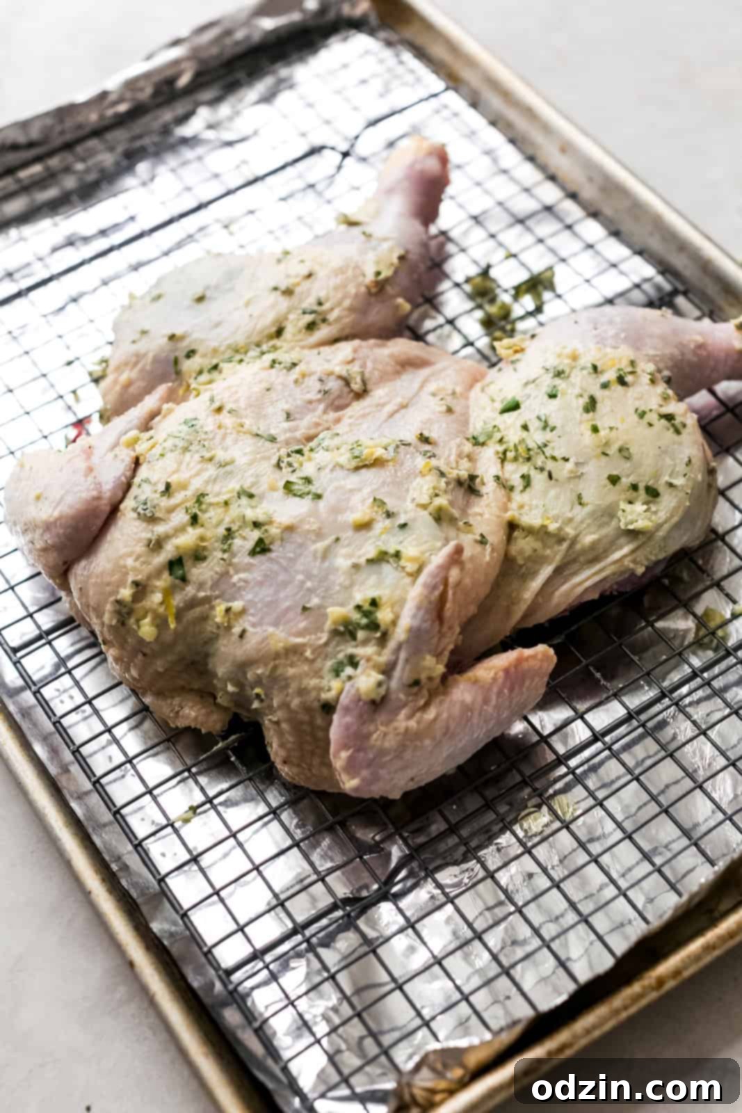 Raw spatchcock chicken generously rubbed with fragrant herb and garlic butter, positioned on a wire rack over a foil-lined baking sheet, ready for roasting.
