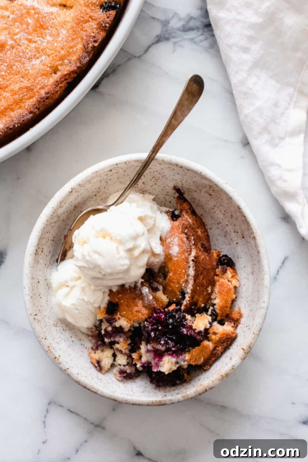 Effortless Texas Blueberry Cobbler 2 blueberry cobbler in speckled bowl with ice cream and spoon on marble