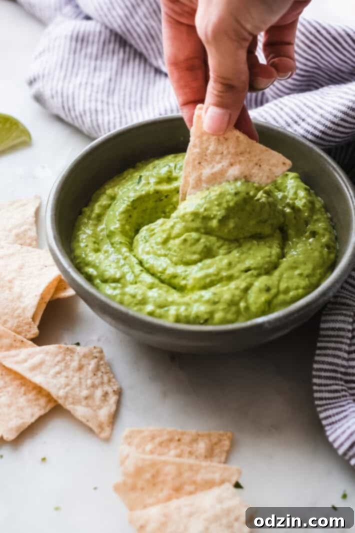 Velvety 5-Minute Avocado Dip 2 Tortilla chip being dipped into a luscious green blender avocado dip served in an olive colored bowl, highlighting its creamy texture.