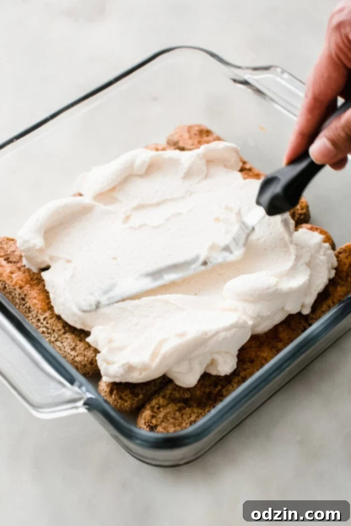Using an offset spatula to neatly spread the creamy mascarpone mixture over a layer of espresso-soaked ladyfingers in a baking dish.