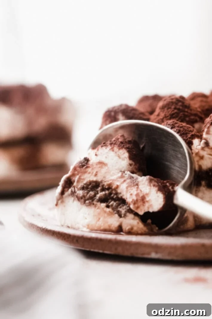 A spoon breaking into a square of eggless tiramisu on a speckled plate, revealing the layers beneath the cocoa dusting.
