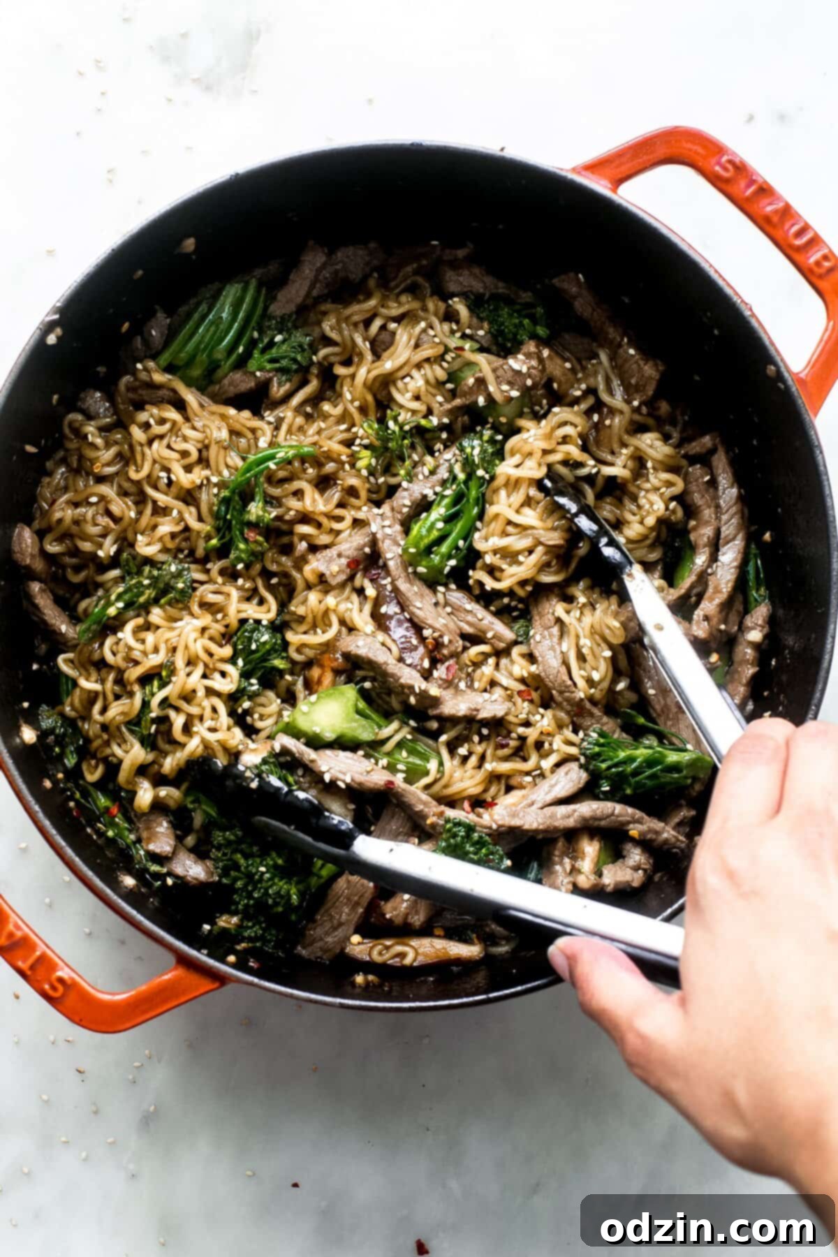 broccoli beef ramen being lifted with tongs from cast iron pot, showcasing the noodles, beef, and broccoli