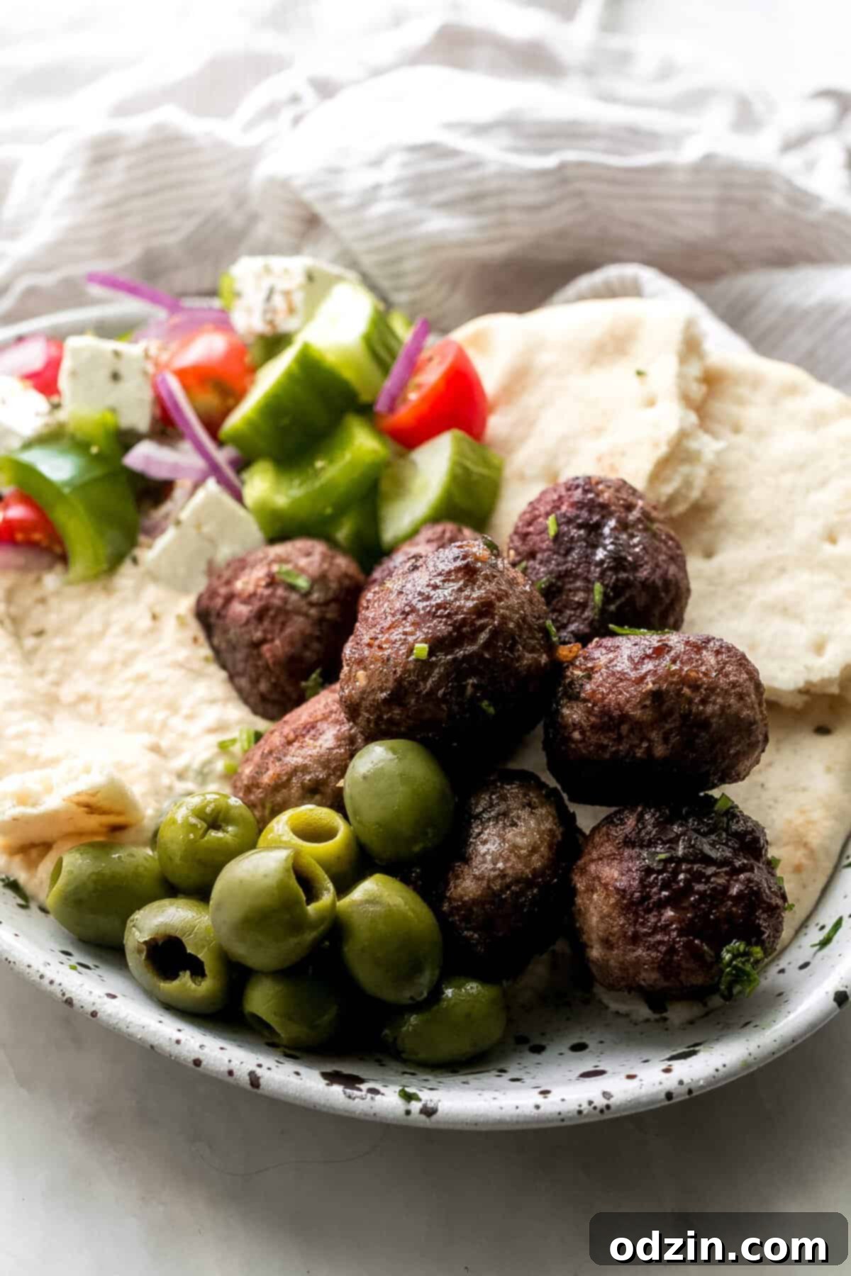 Authentic Greek Keftedes 3 Greek Meatballs (Keftedes) served on a speckled plate with hummus, pita bread, olives, and a fresh Greek salad for a complete mezze meal