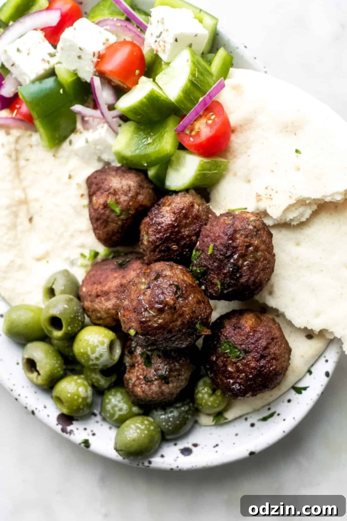 Authentic Greek Keftedes 7 Greek Keftedes meatballs beautifully arranged on a platter with green olives, torn pita bread, a side of Greek salad, and a bowl of hummus
