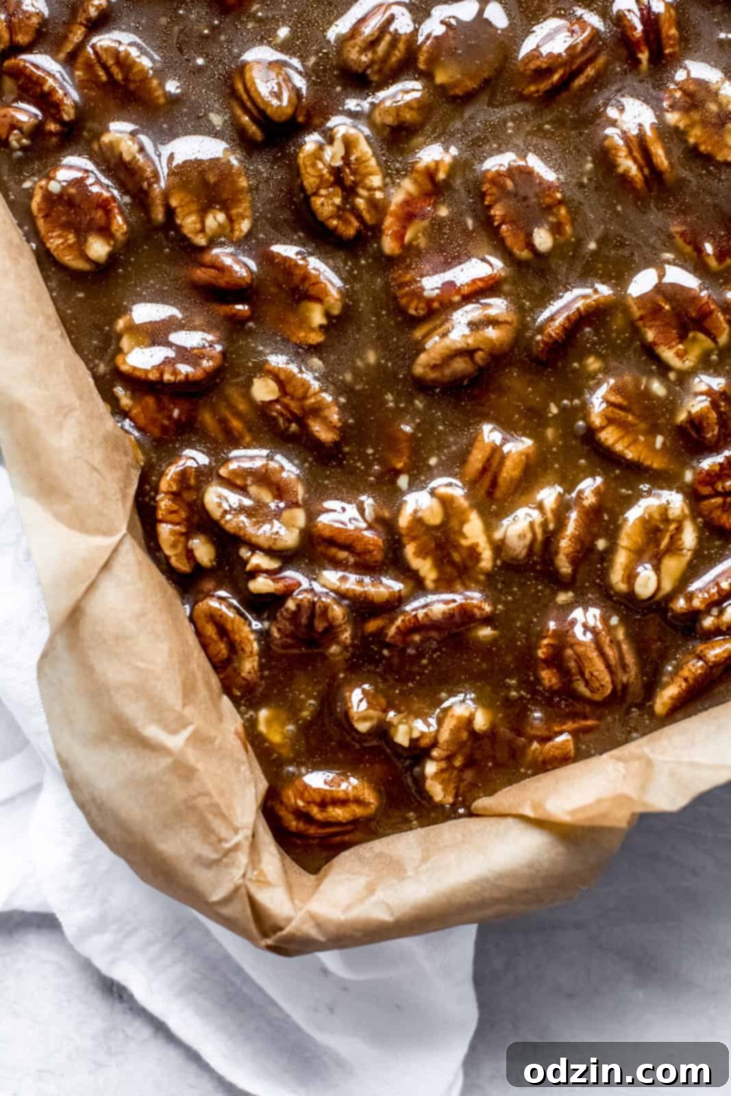 Warm pecan pie topping being poured evenly over the baked cheesecake layer in a parchment-lined baking dish