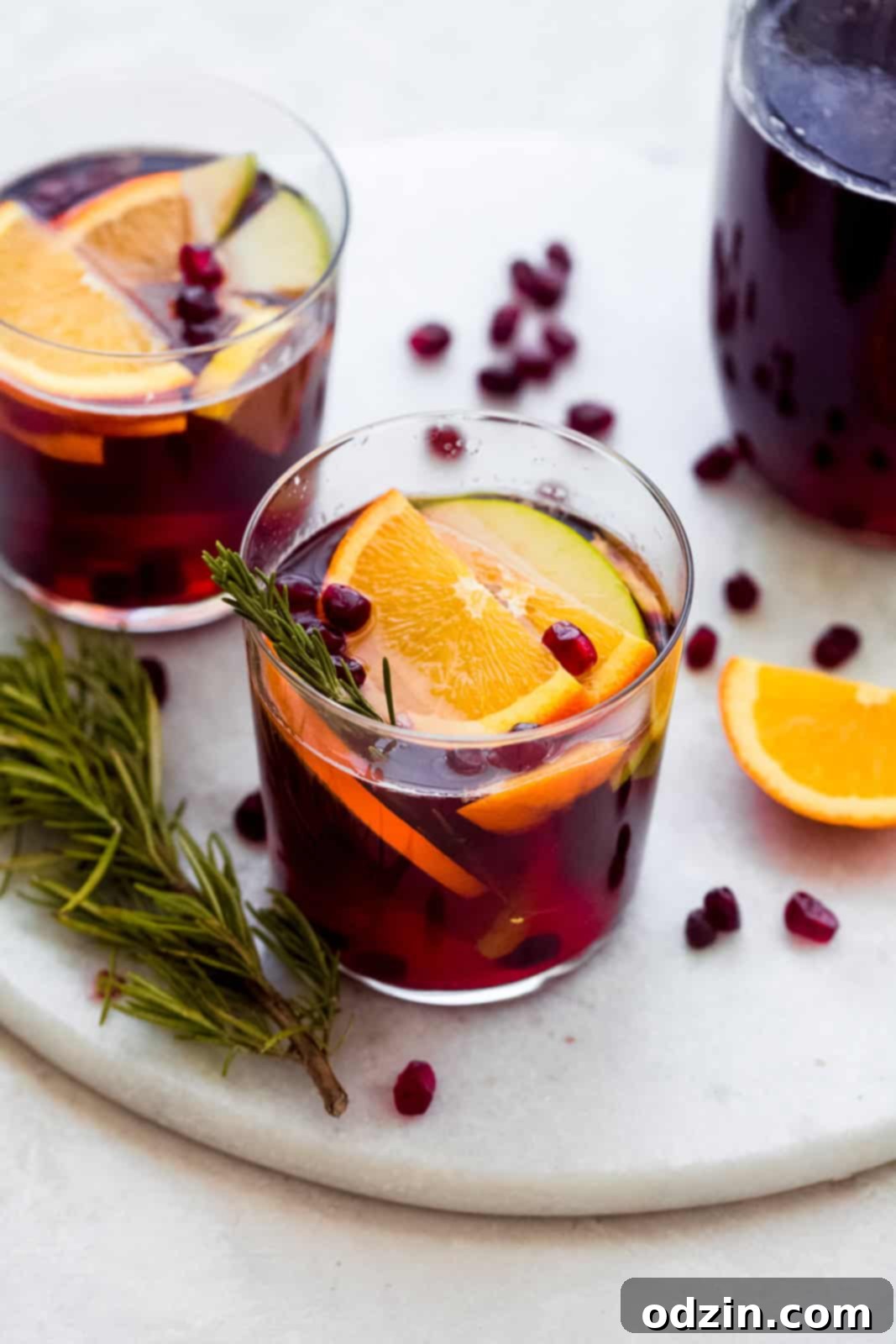 mocktail in glass on marble with oranges, apples, pomegranate arils, and rosemary in glass