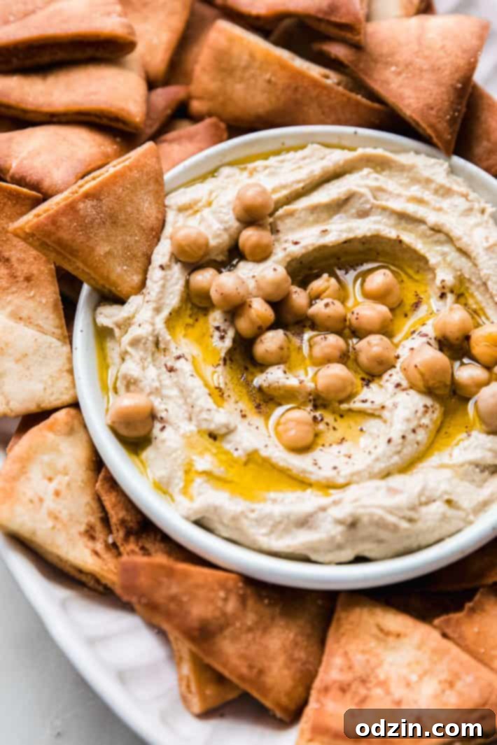 pita chip dipped in homemade hummus A delectable bowl of homemade hummus, beautifully garnished with whole chickpeas and a sprig of fresh herbs, with a pita chip resting invitingly in the dip.