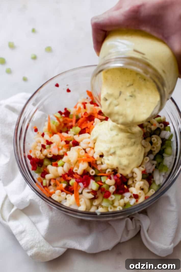 Crave-Worthy Macaroni Salad 7 hand pouring prepared dressing from jar into bowl with pasta and vegetables for salad