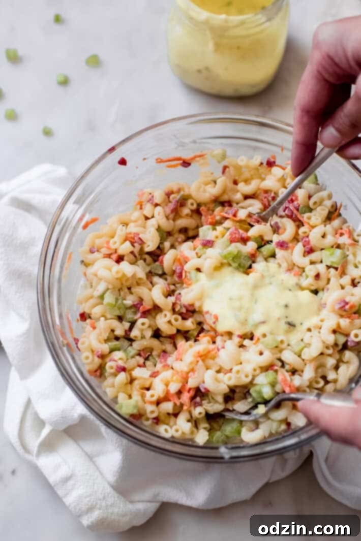 Crave-Worthy Macaroni Salad 9 hand tossing dressing into macaroni salad in glass bowl on white marble
