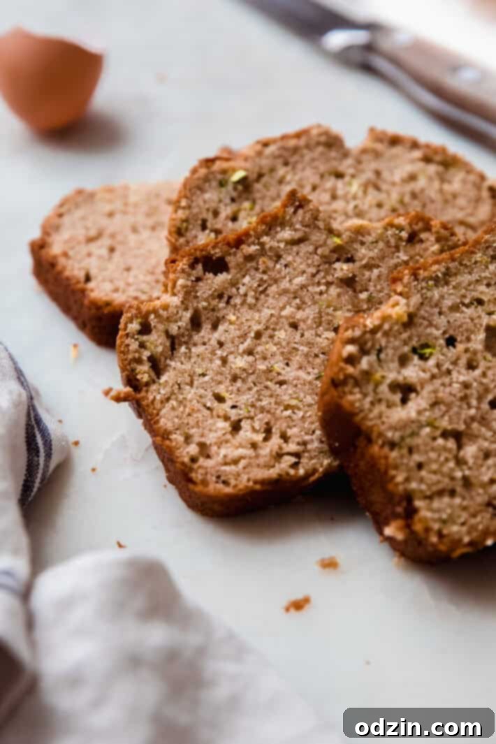 Beautifully arranged slices of zucchini loaf on white marble, with a bread knife, eggshells, and a tea towel, highlighting the texture.