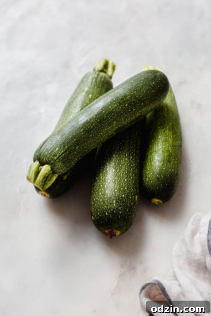 Four fresh zucchini squashes arranged on white marble with a tea towel, ready for grating.