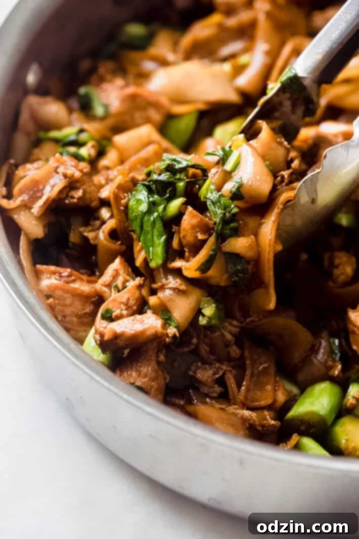Close-up shot of tongs lifting a portion of Pad See Ew noodles, showcasing their glossy coating and ingredients.