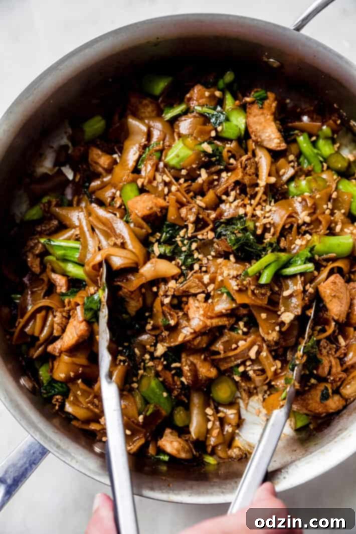 A pair of tongs expertly tossing Thai noodle stir-fry in a hot skillet, showcasing the cooking process.
