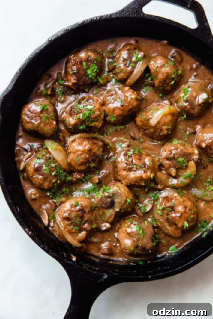 Golden brown Salisbury steak meatballs simmering in a rich, dark mushroom and onion gravy in a cast iron skillet on a white marble surface.