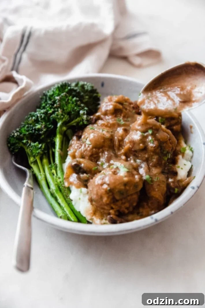 A comforting bowl of mashed potatoes topped with Salisbury steak meatballs and a side of sautéed broccolini, garnished with fresh herbs.