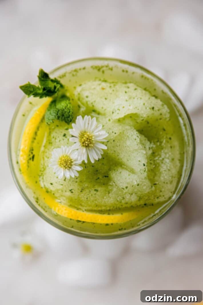 Close-up of a glass filled with vibrant green blended mint lemonade slushie, topped with a mint sprig, on a white marble surface.