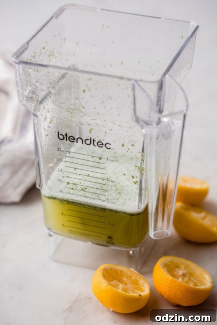 A blender filled with a vibrant green mixture of lemon juice, lime juice, fresh mint, sugar, and water, ready for ice, on a white marble surface.