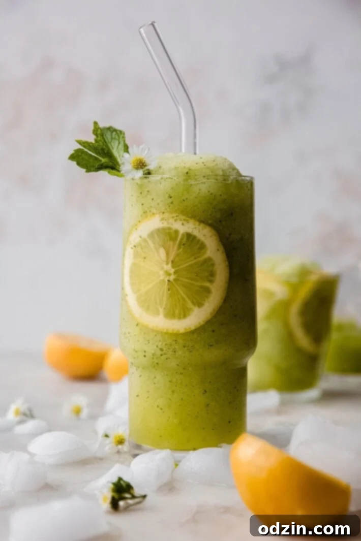 A tall glass of vibrant green frozen mint lemonade with a glass straw and lemon slice, showcasing its frosty texture.