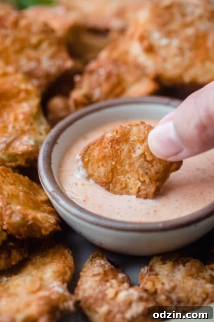 Close-up shot of a single golden-brown air fryer fried pickle being dipped into a creamy, vibrant remoulade sauce.
