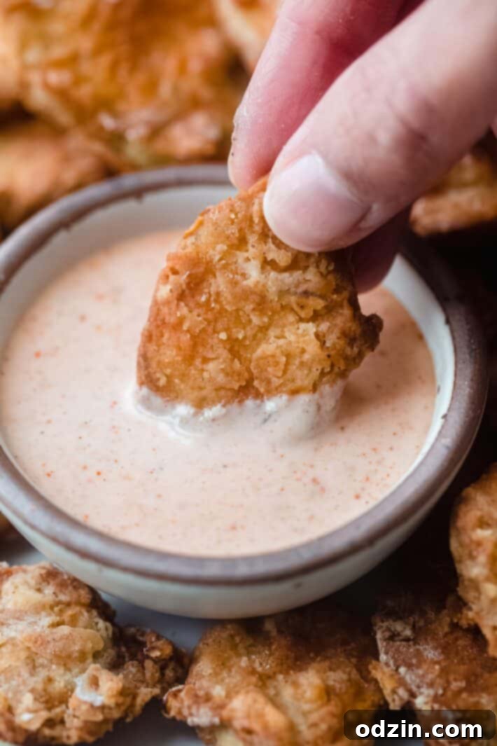 A hand reaching into a bowl to dip a golden-brown fried pickle into a creamy remoulade sauce.