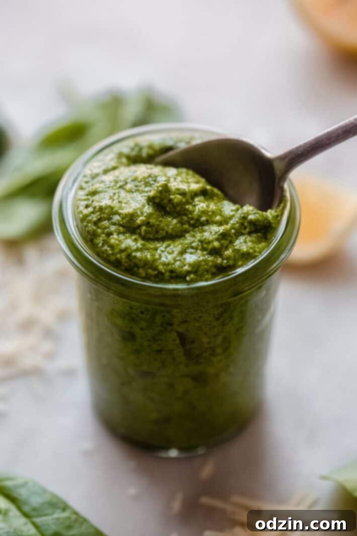 A wide shot of a glass jar filled with green pesto, surrounded by its fresh ingredients like vibrant spinach, crunchy walnuts, garlic, and bright lemons on a rustic wooden surface.