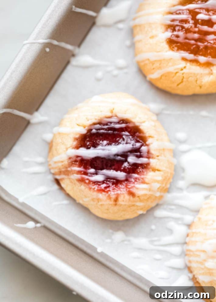 A perfectly baked thumbprint cookie, topped with a delicate white glaze and a dollop of raspberry jam in its center.