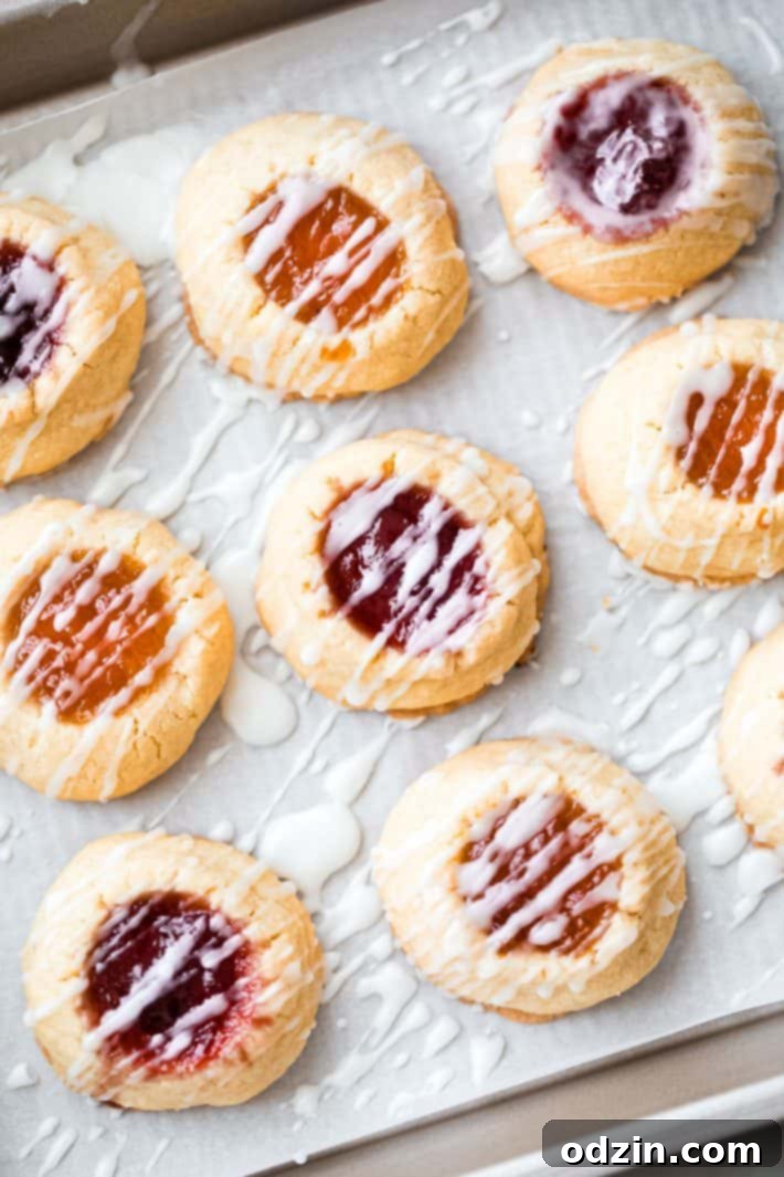 A baking sheet filled with glazed, jewel-toned thumbprint cookies, showcasing the vibrant jam and delicate white drizzle.
