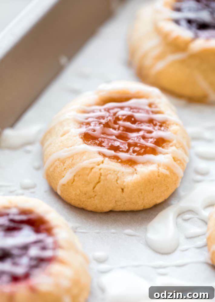 A close-up of a single thumbprint cookie, showcasing its golden edge, creamy glaze, and bright apricot preserves filling.