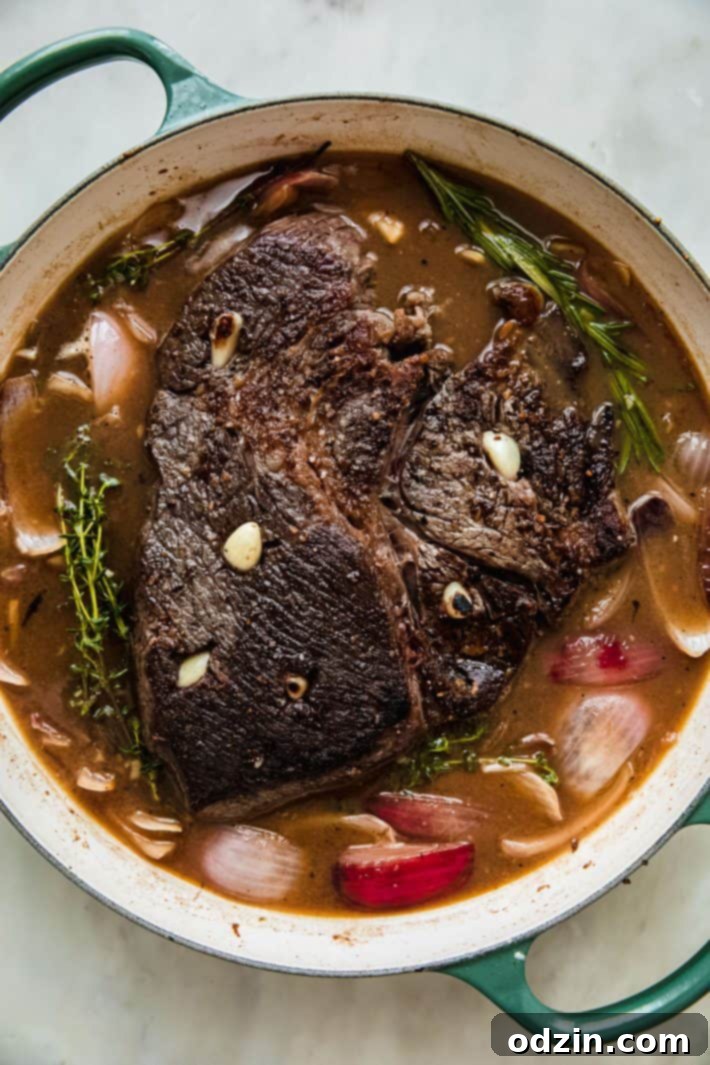 A dutch oven filled with a seasoned pot roast, quartered onions, whole garlic cloves, and fresh thyme, ready for braising.