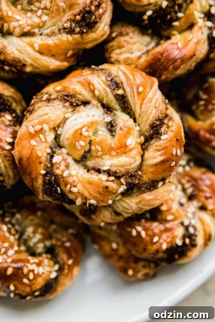 Close-up shot of a single golden-brown za'atar puff pastry swirl on a white plate, highlighting its intricate layers, perfectly formed spiral, and delicious sesame seed topping.