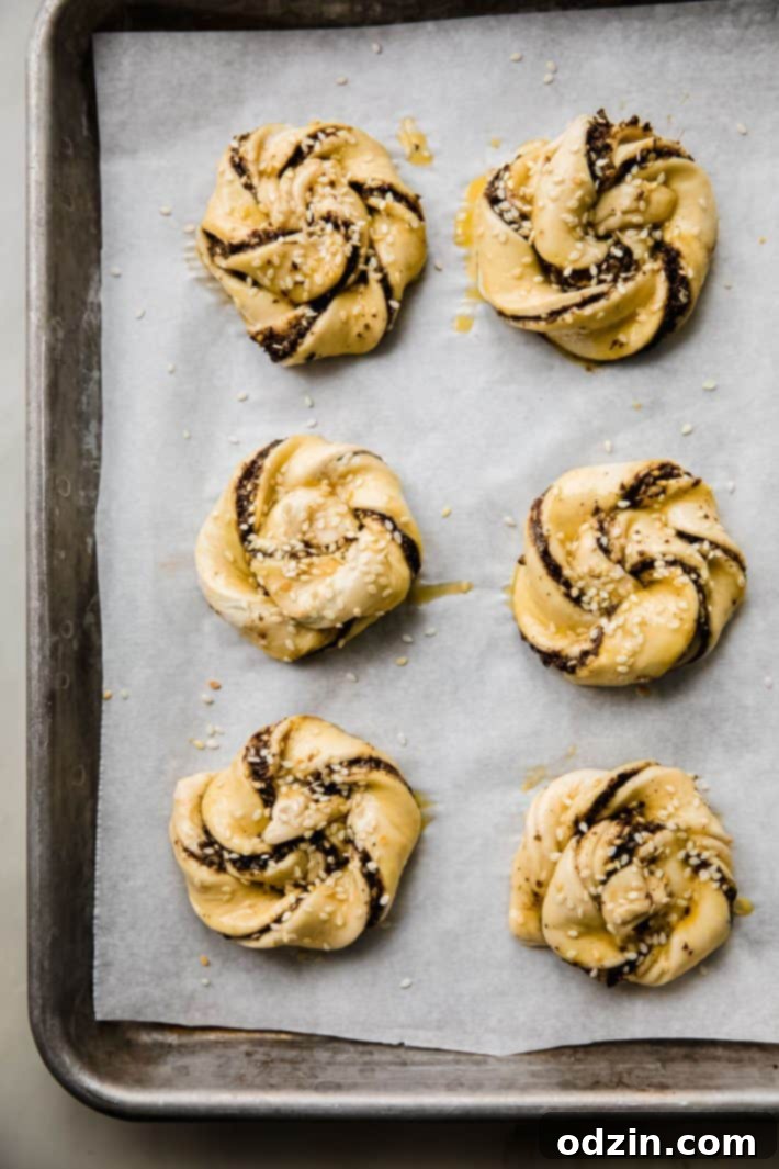 Puff pastry swirls neatly arranged on a sheet pan, already brushed with egg wash and topped with sesame seeds, poised and ready for baking in a hot oven.