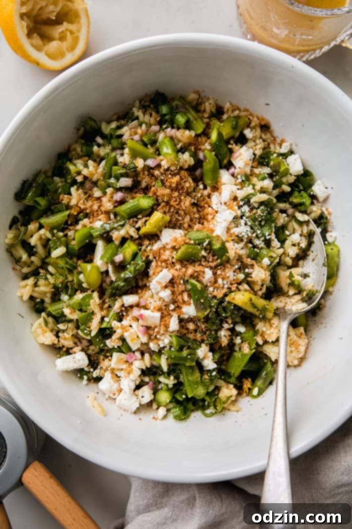 Fresh and vibrant asparagus orzo salad in a bowl with a serving spoon, garnished with herbs and feta