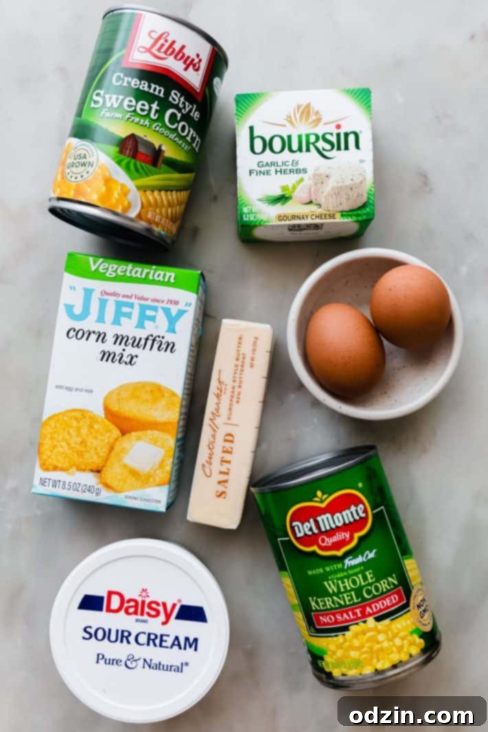 Assortment of ingredients for Jiffy Corn Casserole, including Jiffy mix, cans of corn, eggs, butter, sour cream, and a puck of Boursin cheese.